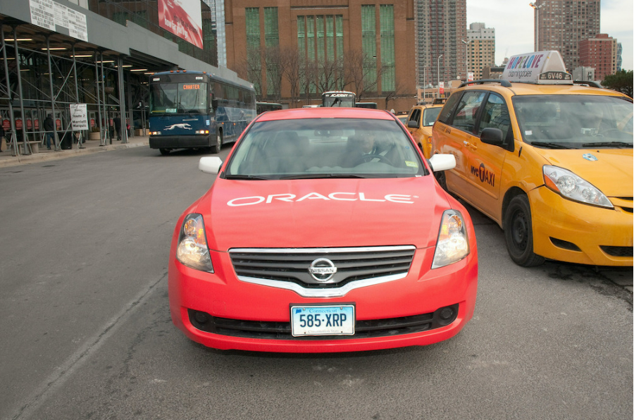 Imagen de un automóvil que conduce por una carretera, con Oracle escrito en él. A un lado hay un autobús, al otro un taxi.
